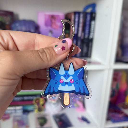 Hand holding a blue cartoon gargoyle character keychain in front of a colorful shelf.